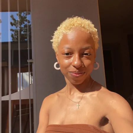 Carly is looking at the camera wearing a tan tube top, a silver cross necklace, and white hoop earrings