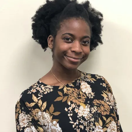 Khangelani is sitting, looking at the camera, and smiling. She is wearing a black top with a floral design and a gold necklace. Her hair is pulled up with a braid down the middle.
