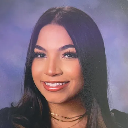 Ana is smiling at the camera in front of a purple background. She is wearing a dark colored top and her hair is parted down the middle.