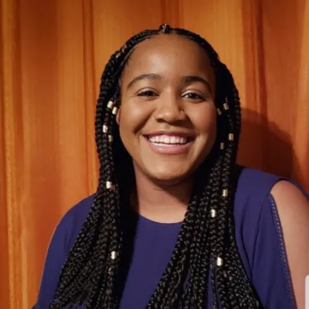 Letícia is in front of a wooden wall smiling at the camera. She is wearing a purple blouse and her braided hair has gold hair jewelry in it.