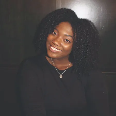 Trinity is sitting in front of a dark wood wall smiling at the camera. She is wearing a black blouse and her curly hair is parted down the middle.Trinity is sitting in front of a dark wood wall smiling at the camera. She is wearing a black blouse and her curly hair is parted down the middle.Trinity is sitting in front of a dark wood wall smiling at the camera. She is wearing a black blouse and her curly hair is parted down the middle.