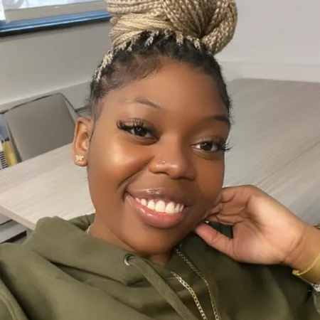 Thais is smiling at the camera in a classroom. She is wearing a green hoodie and her braids are in a high bun.