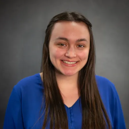 Gabrielle is sitting in front of a grey background and smiling at the camera. She is wearing a blue blouse and her long, brown hair is parted down the middle.