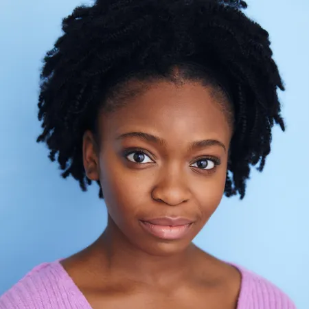 Nicole-Bethany is looking at the camera in front of a blue background. She is wearing a pink sweater and her hair is pulled up into a pony tail.