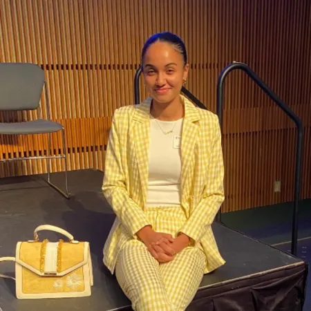 Quincie is sitting on a stage in a yellow gingham suit, smiling.