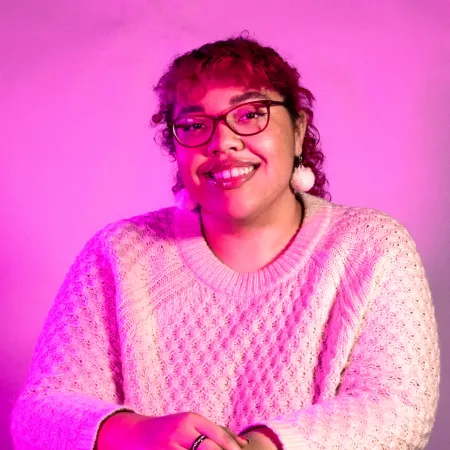 Avery is smiling at the camera. They are wearing a white sweater, dangling puff earrings, and glasses. The photo is tinged pink.