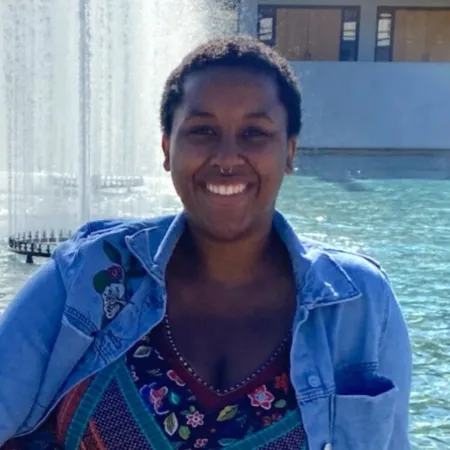 Kyla is standing in front of the IC Fountains smiling at the camera. She is wearing a jean jacket and a multicolored shirt. She has a gold hoop septum piercing.
