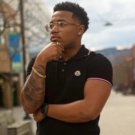 Malachi is looking off in the distance with his hand on his chin. He is wearing a black shirt, glasses, and a gold bracelet 