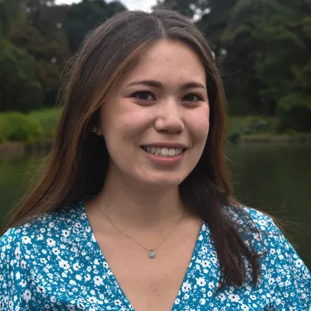 Jessica is looking at the camera and smiling. She is outdoors, in front of a body of water. She is wearing a blue floral top and her hair is parted down the middle.