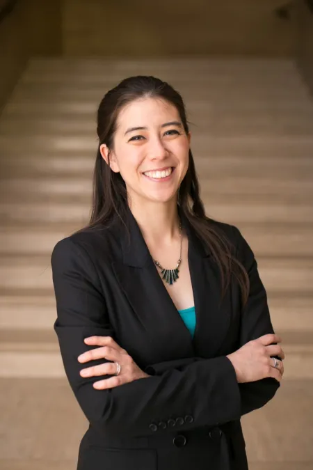 A smiling woman with long brown hair, wearing a blazer, with her arms crossed over her chest.