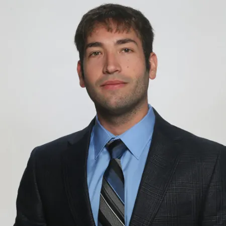 Headshot image of Dan DeCaria on white background. He is wearing a navy blue patterned suit coat over a light blue dress shirt and patterned blue and grey striped tie. He is facing the camera directly with expressionless facial posture.