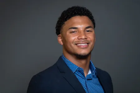Adrian is posing for photo on dark grey background. Is wearing royal blue shirt with white dots and a dark blue blazer. Is clean shaven and has short black hair. Is smiling.