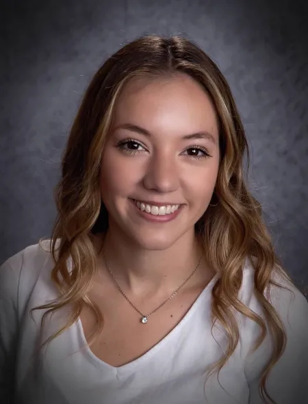 Alissa is posing in front of dark grey background. Is wearing white top and necklace. Has light blonde hair extending past shoulders. Is looking at camera and smiling.
