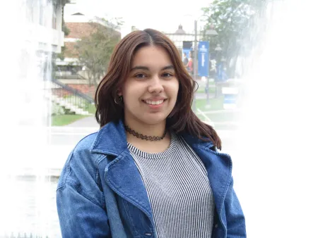 Ana is posing for picture with Ithaca College academic row in background. Is wearing a grey shirt with a blue jacket. Has brown hair extending to shoulders. Is looking at the camera and smiling.