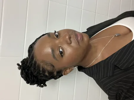 Azaria is posing on white brick background. Is wearing white top with black blazer with thin white pinstripes. Is wearing a necklace. Has braided hair pulled back into bun.