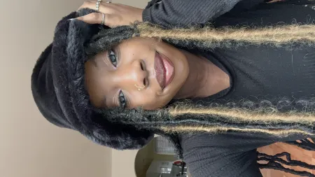 Caleigh is posing for picture with beige background. Is wearing a black shirt and black fur hat. Has braided hair dyed blonde on the end and dark roots. Hair is long extending past torso.