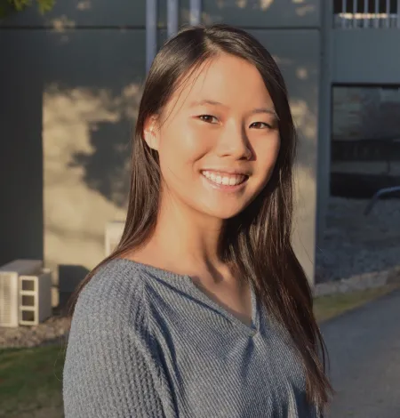 Emily is posing with building in background. Is wearing a grey shirt and has long dark hair extending past shoulders. Is smiling.