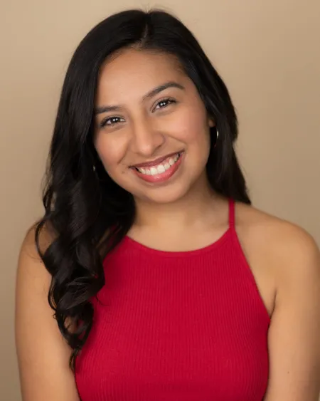Erica is posing for a standard headshot on beige background. Is wearing a red top with exposed shoulders. Is smiling.