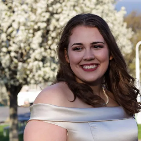 Jeannette is posing for photo with white blooming tree in background. Is wearing white dress with exposed shoulders. Has a pearl necklace. Is looking into the distance  to the left of the camera and smiling. Has dark shoulder-length hair.