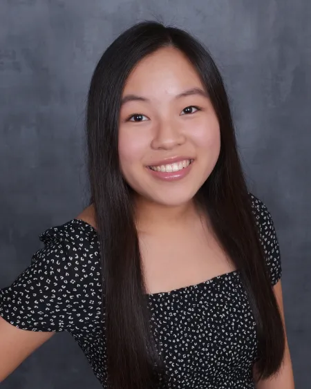 Jillian is posing for standard headshot on charcoal grey background. She is wearing a black top with white spots and has long black hair extending past shoulders. She is smiling and looking directly at the camera.