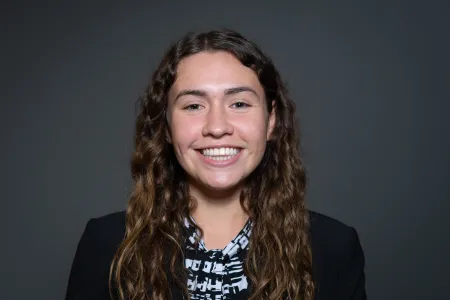 Julie is posing for photo on dark grey background. Is wearing blue and white patterned shirt with blue blazer. Has long brown hair extending past shoulders. Is looking at camera and smiling.