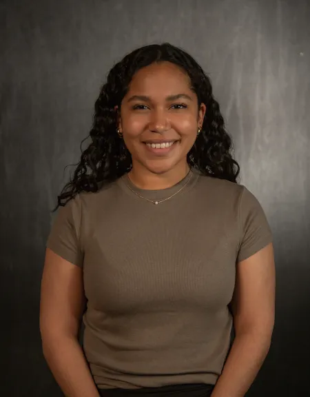 Leanna is posing on grey patterned background. Is wearing light brown shirt and necklace. Has long black hair extending past shoulders. Is looking at camera and smiling.