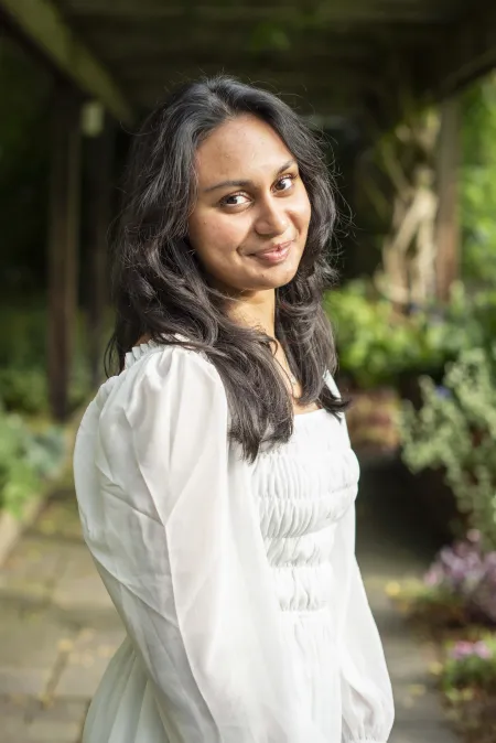 Liza is posing with green plants in background. Is wearing white top. Has dark hair extending to shoulders.