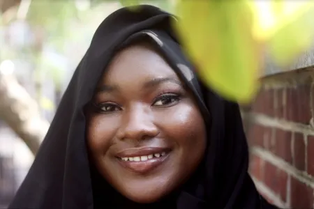 Makiyah is posing for photo with green leaf and brick facade in background. Is wearing black. Staring at the camera and smiling.