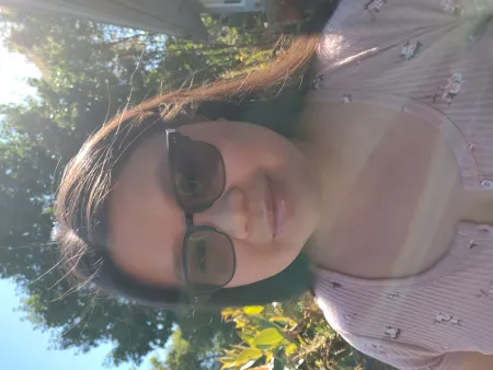 Miriam is posing in front of trees in background on sunny day. Is wearing a light pink patterned top and sunglasses. Has dark brown hair shoulder length.