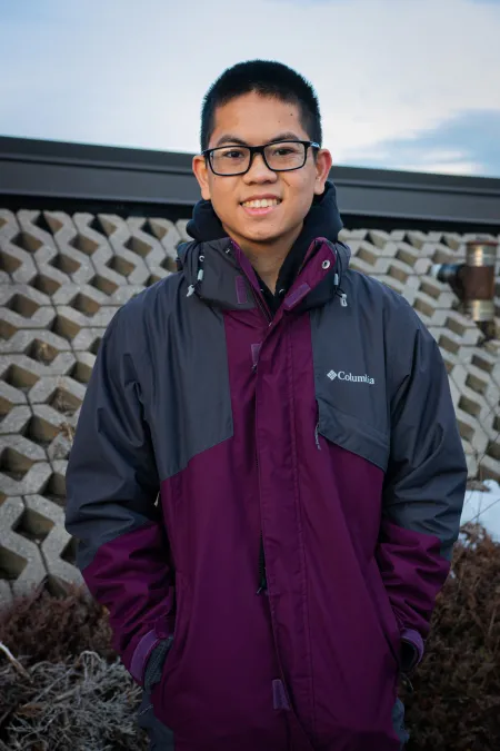 Nicholas is posing for picture in front of patterned brick facade.  Is wearing a grey and purple coat and glasses. Is looking at the camera and smiling.