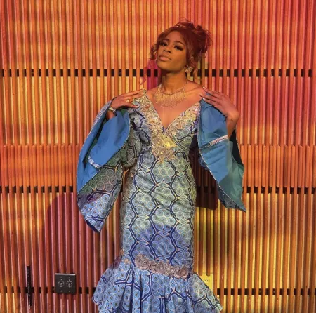 Is wearing a long blue and seafoam green dress with extended sleeves. Is posing on orange patterned background. Looking at camera, but not smiling.
