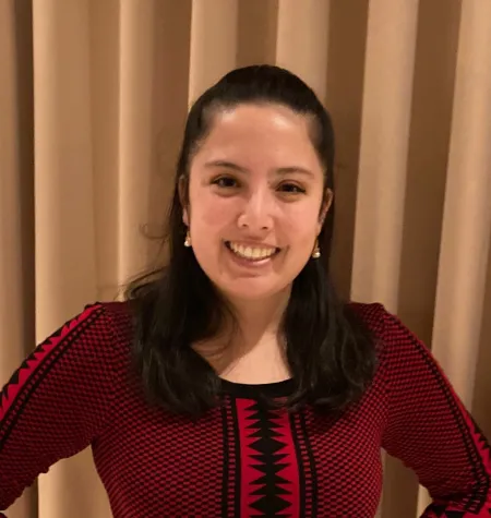Rebecca is posing for photo in front of beige curtains. Is wearing a maroon top with red and black pattern stripes. Has dark hair extending past shoulders. Is looking at camera and smiling.