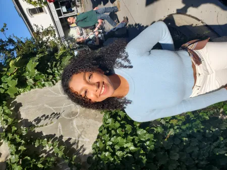 Stephanie is posing for photo in front of brick wall covered in green ivy. Serindipity building is in the background as well as two bypassers. Stephanie is wearing light blue sweater and white pants. Has dark curly hair extending to shoulders. Is looking at camera and smiling.