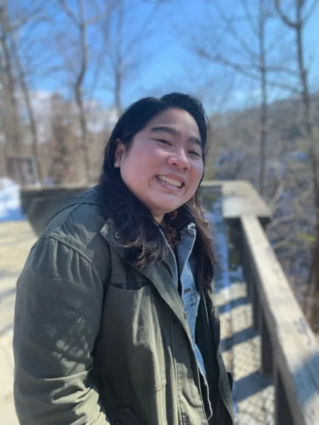 Violet is posing outside on cold day with barren trees in background. Is wearing a olive green winter coat. Has shoulder length dark hair.