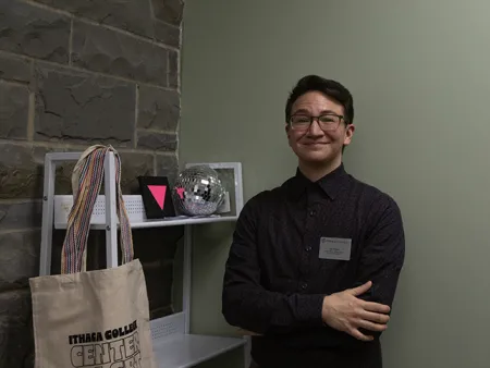 Pride Fellow Lee Tyson, a masculine half-East Asian person with short brown hair and glasses, stands next to a bookcase that holds a shiny disco ball and a painting of a pink triangle against a black background.