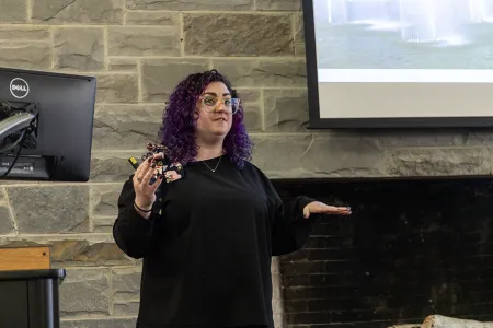 LGBTQ Center Director Crissi Dalfonzo, a woman with curly purple hair, glasses, and a dark shirt, stands next to a project and podium, speaking to a room.