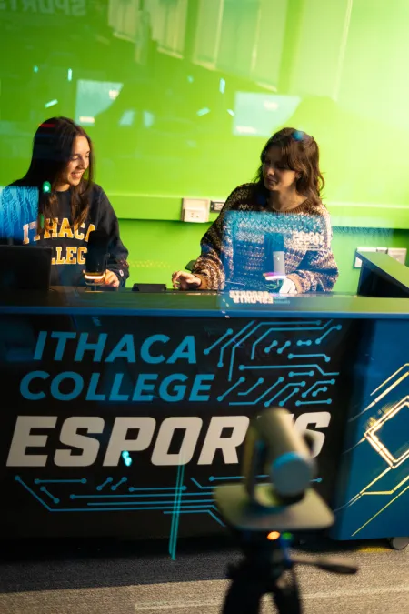 Two students in front of a greenscreen in a broadcast booth labeled "Ithaca College Esports."