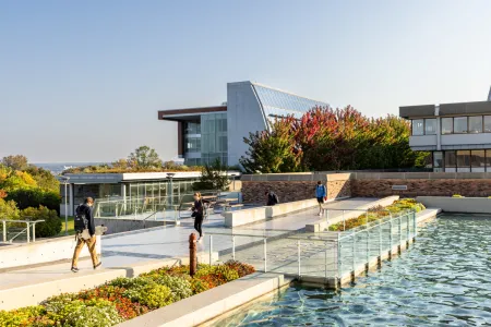 Students walk on a college campus with a fountain pool in the foreground and a modern glass academic building in the background. 