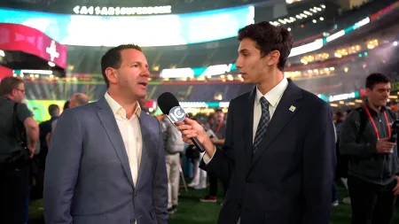 A college student holds a microphone as he interviews a man at an NFL football field. 