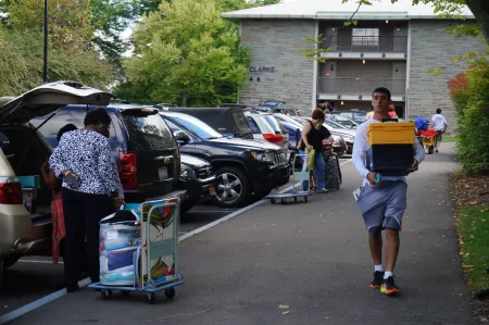 students and parents carry cases and boxes along a sidewalk outside a residence hall