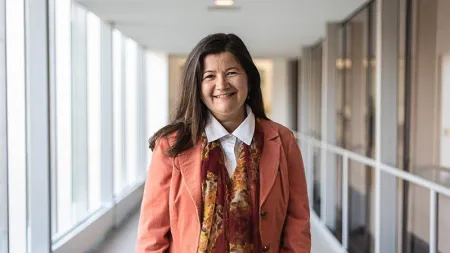 Headshot of Ana Paula Pimentel Walker. Ana Paula is wearing a salmon suit coat with a white collared shirt. Is posed in a long hallway.