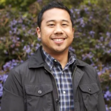 Headshot of Eric Pido. Eric is wearing a grey jacked with chest pockets and a blue and white flannel. He is posed in front of a flowering bush.