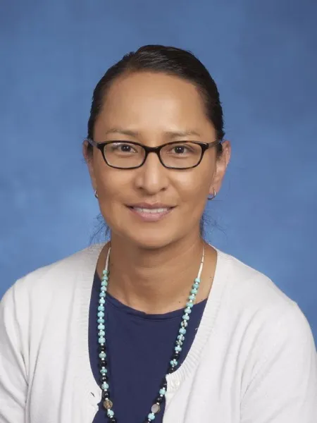 Headshot of Hollie Kulago. Hollie is wearing a white cardigan and blue shirt. She is also wearing a beaded light blue necklace and glasses.