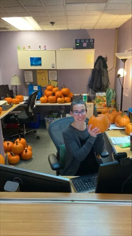 Picture of Marney holding a pumpkin, smiling at the camera, while seated at her desk.