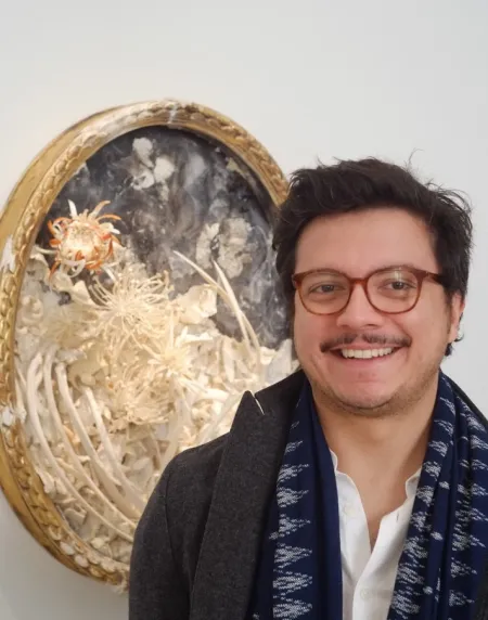 Headshot of Josh Franco. Josh is wearing a charcoal grey peacoat and blue/white pattered scarf, with a white buttoned shirt. Is posed in front of a painting.