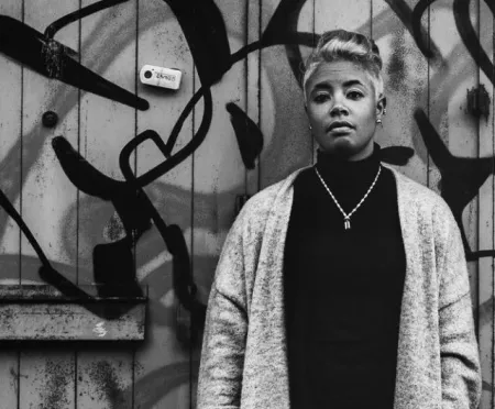 Headshot of Lillian-Yvonne Bertram. Lillian-Yvonne is posed in front of patterned background. Is wearing a black sweater with white/grey cardigan.