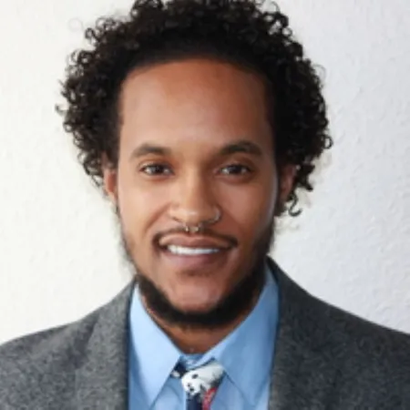 Headshot Shahan Bellamy. Shahan is wearing a grey suit and light blue collared shirt and patterned tie.