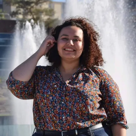 Half-length portrait of Geselle from waist up, smiling and standing in front of the Dillingham fountains. Geselle is wearing a colorful floral button-down shirt tucked in jeans, and her left arm is raised in mid-motion, tucking hair behind her ear as she faces the camera.