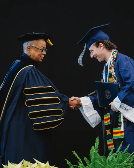 A woman wearing academic regalia shakes hands with a graduate.