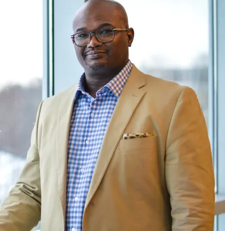Man standing in front of a window wearing a sport coat and blue checked shirt.